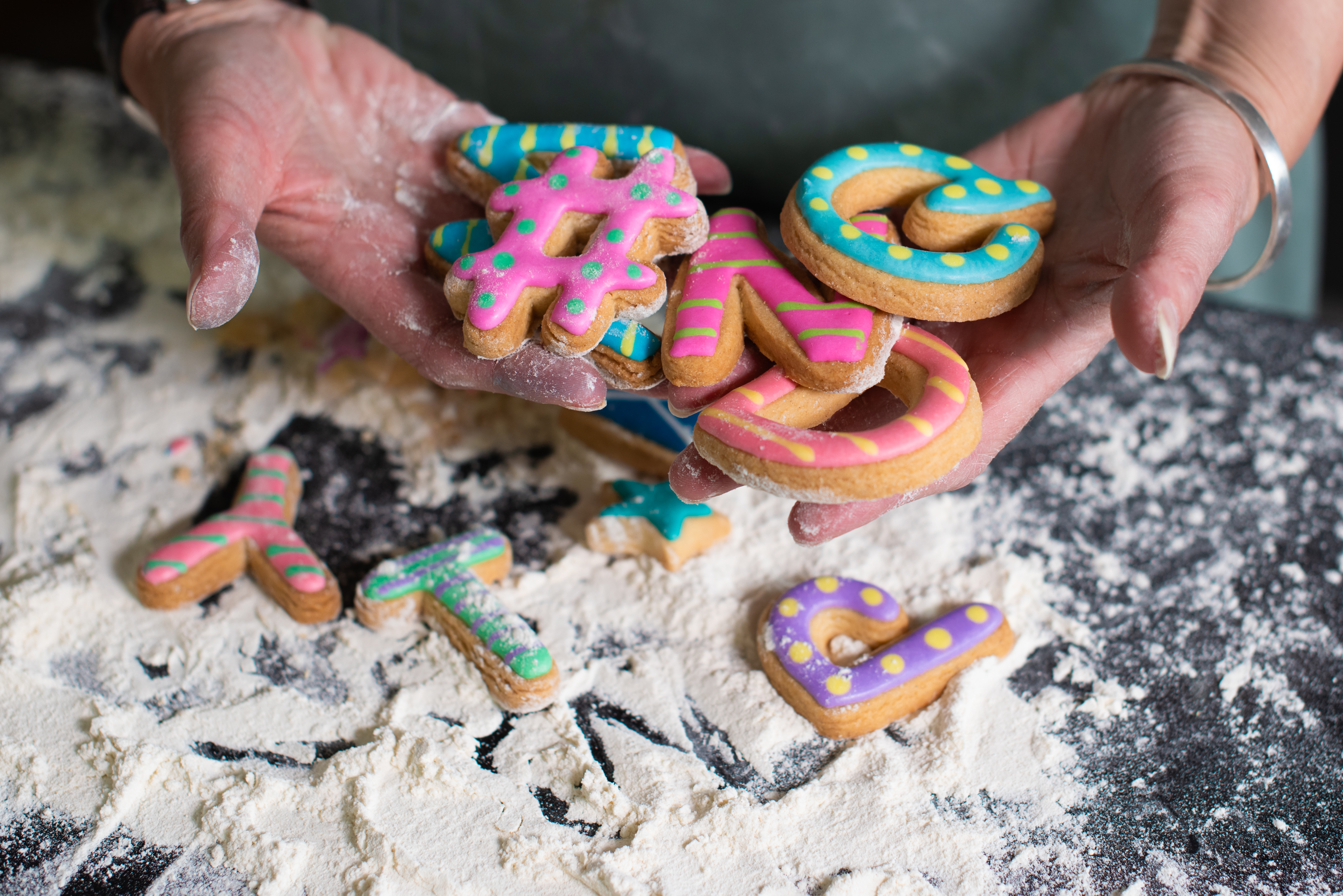 A close-up of hands holding colorful, decorated cookies shaped like letters and symbols, including a hashtag and an at symbol. The cookies are iced in bright colors with various patterns. The countertop below is covered in flour, with more cookies scattered around. The hands and the cookies have traces of flour on them.